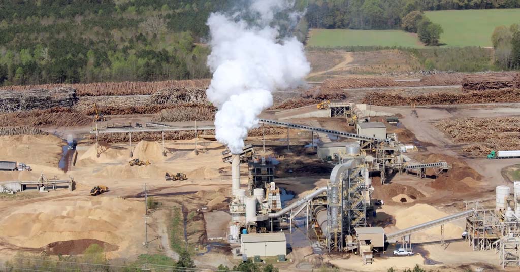 Aerial view of a wood pellet plant with a tall steam plume and piles of sawdust